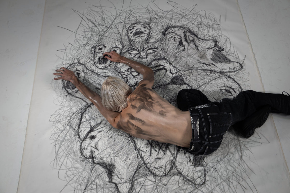 ONEFATE Campaign 01, model in black jeans lying across a large charcoal drawing of faces, seen from above