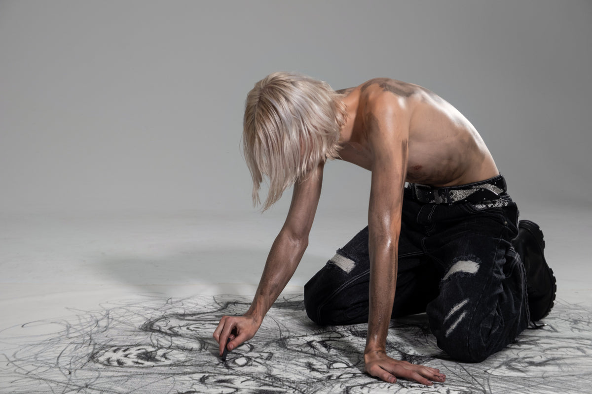 ONEFATE Campaign 01 – blond model kneeling and drawing with charcoal on the floor, wearing snake belt and snake denim