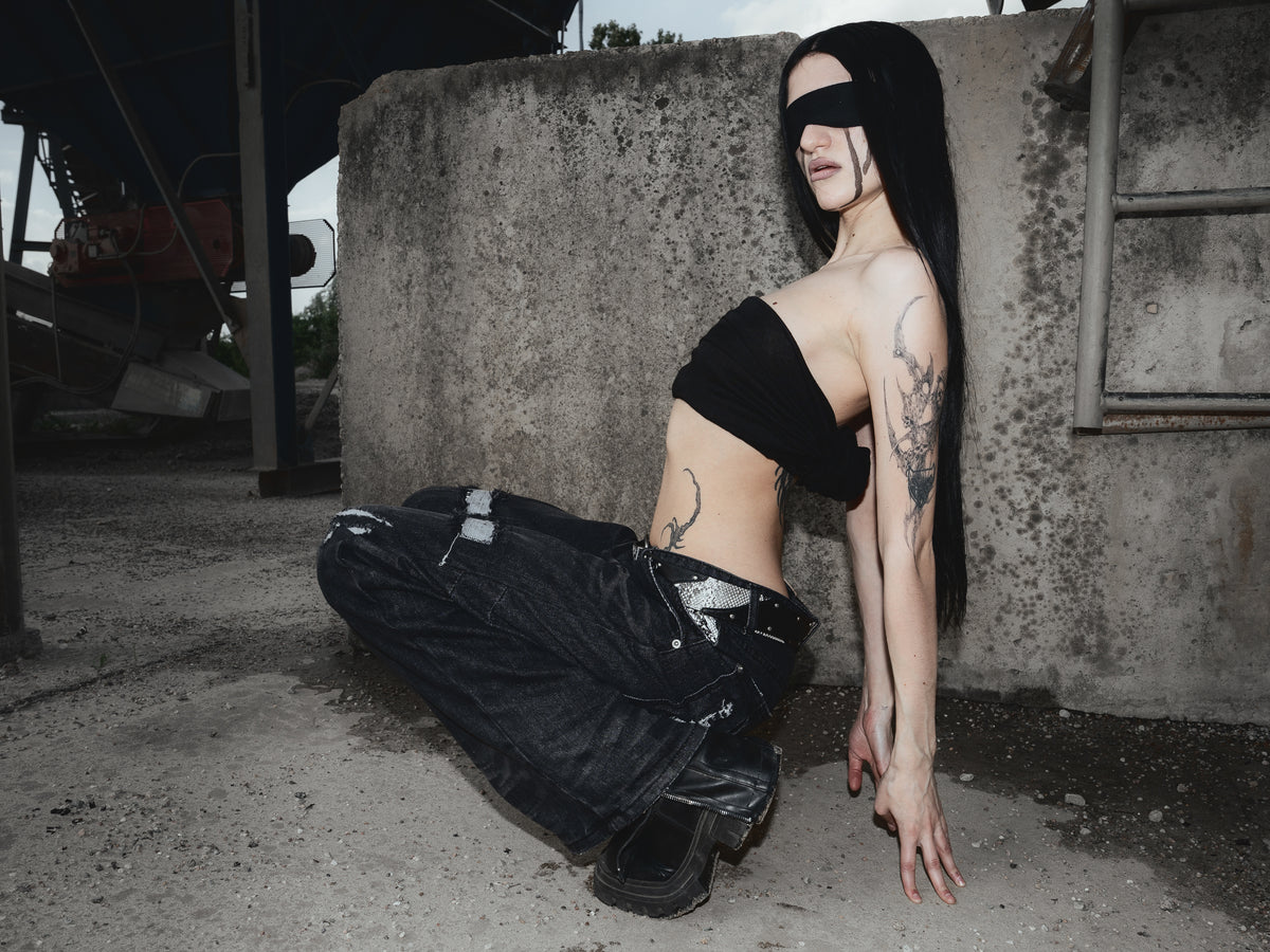 ONEFATE Campaign 02 – blindfolded model crouching at a concrete block in black snake denim and snake belt